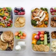 Four clear containers are filled with a variety of foods, including sliced vegetables, fruits, boiled eggs, nuts, crackers, grilled chicken, and salads. Each container has multiple compartments, separating different food items for a balanced meal prep. 30 Easy and Tasty Healthy Food Ideas for Choosy Eaters