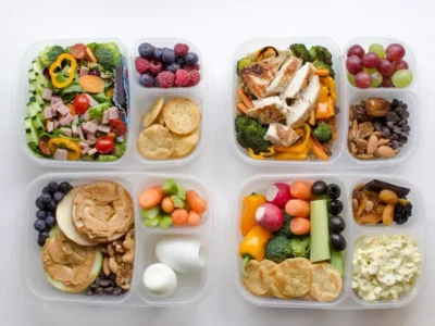 Four clear containers are filled with a variety of foods, including sliced vegetables, fruits, boiled eggs, nuts, crackers, grilled chicken, and salads. Each container has multiple compartments, separating different food items for a balanced meal prep. 30 Easy and Tasty Healthy Food Ideas for Choosy Eaters