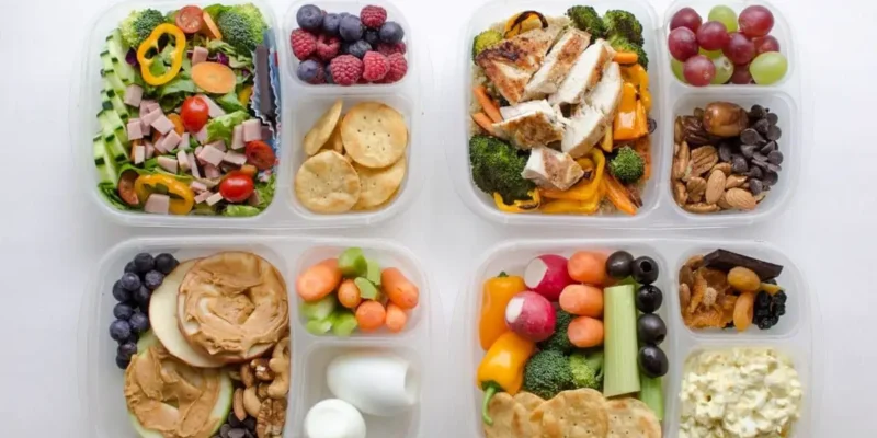 Four clear containers are filled with a variety of foods, including sliced vegetables, fruits, boiled eggs, nuts, crackers, grilled chicken, and salads. Each container has multiple compartments, separating different food items for a balanced meal prep. 30 Easy and Tasty Healthy Food Ideas for Choosy Eaters