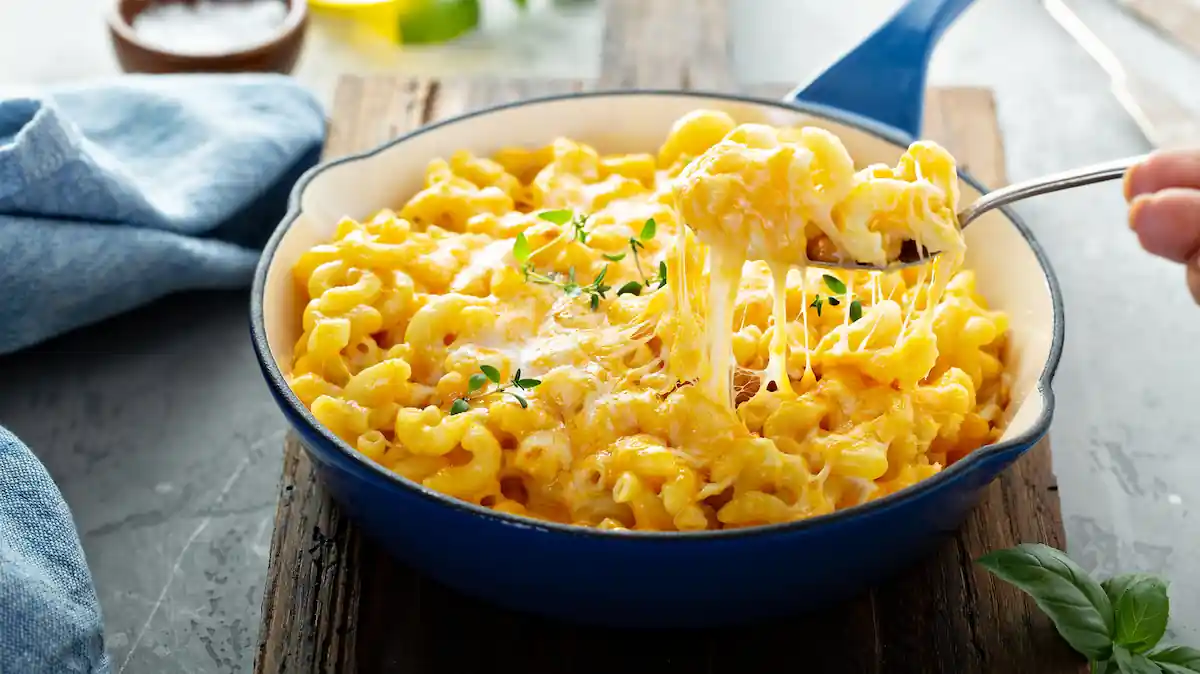 A close-up of a skillet filled with creamy macaroni and cheese garnished with herbs. A person's hand is lifting a forkful of the cheesy pasta, with strings of melted cheese visible. Fresh herbs and a cloth napkin are also in the background. 