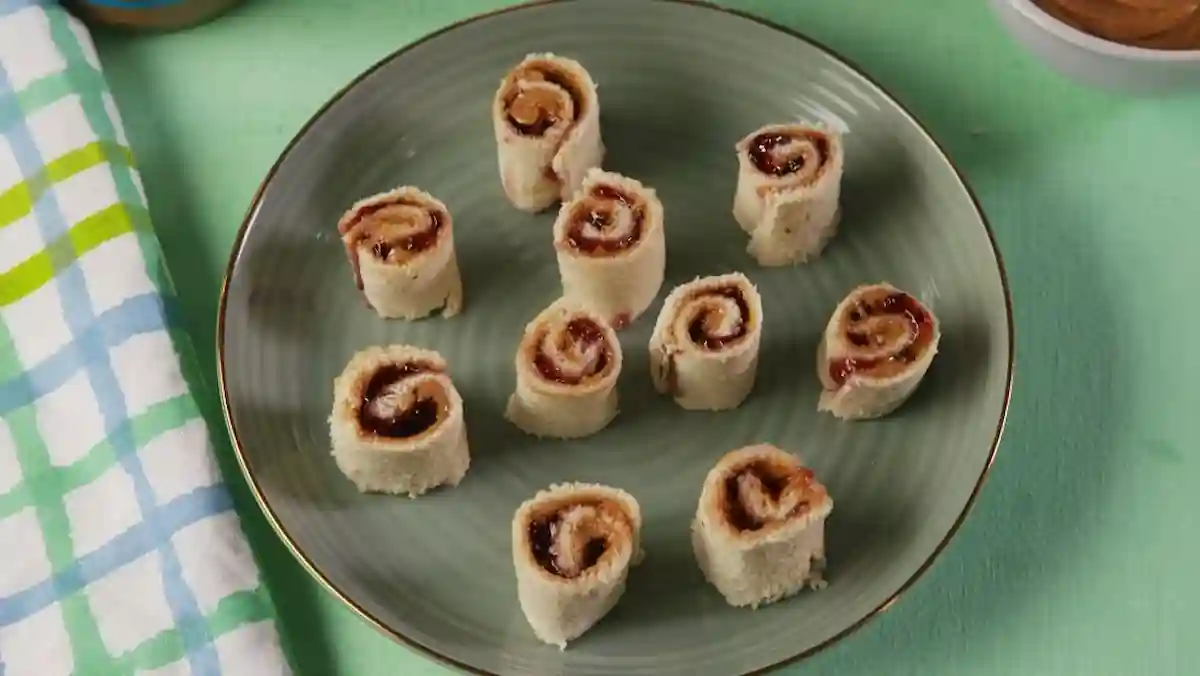 A green plate holds nine small rolls made from bread and filled with a spread, arranged in a circular pattern. The spread appears to be a mixture of peanut butter and jelly, indicated by the brown and dark red swirls within each roll. A green tablecloth is visible underneath.