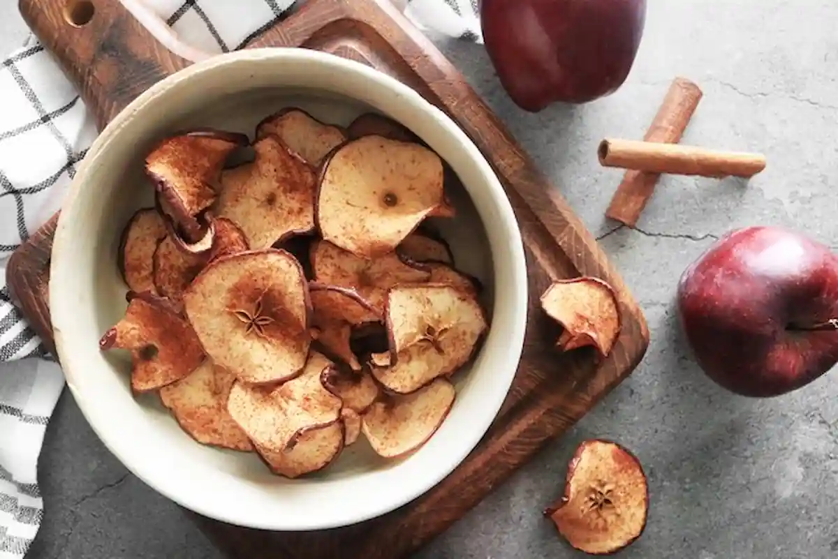 A bowl of homemade apple chips sits on a wooden cutting board with a checkered cloth underneath. Two whole red apples and a cinnamon stick are placed nearby on a gray surface. Some apple chips are scattered around the bowl.