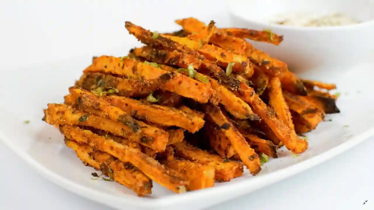 A plate of crispy sweet potato fries seasoned with herbs and spices is placed on a white plate. The fries have a golden-brown appearance and some green garnish. A small cup of dipping sauce is partially visible in the background.