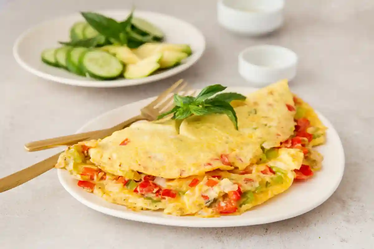 A colorful vegetable omelette folded in half, garnished with fresh basil, is served on a white plate with a fork and knife. In the background, there's a side dish of sliced cucumber and avocado, also on a white plate. Two small empty bowls are placed nearby.