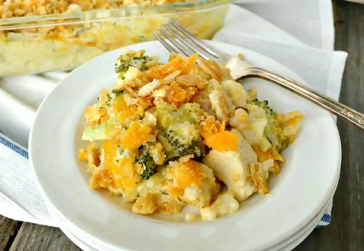 A white plate holds a serving of chicken and broccoli casserole topped with melted cheese and crushed crackers. There's a fork on the plate and a casserole dish in the background on a white cloth.