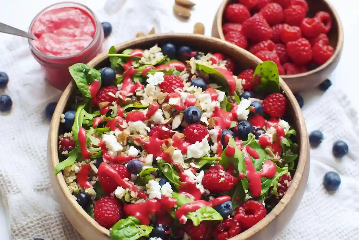 A colorful, fresh salad is served in a bowl, featuring a mix of leafy greens, raspberries, blueberries, crumbled feta cheese, and chopped nuts. The salad is drizzled with a vibrant raspberry dressing. Additional raspberries and a jar of dressing are seen in the background.