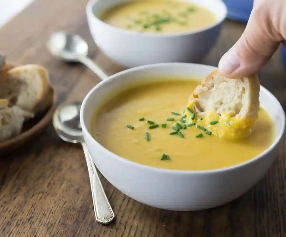 A close-up of a person dipping a piece of bread into a bowl of creamy yellow soup garnished with chopped chives. Another bowl of soup and a plate of bread slices are seen in the background. Two spoons rest on the wooden table. 