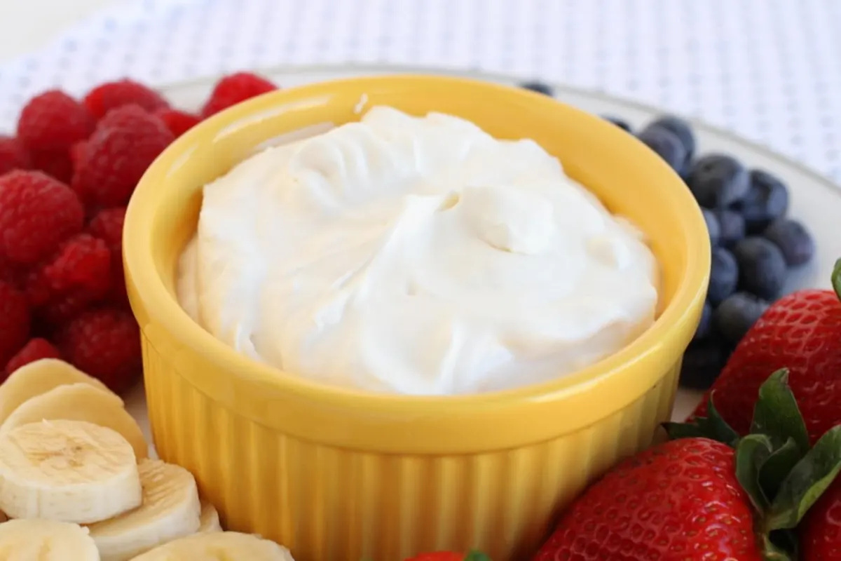 A yellow ramekin filled with creamy dip is surrounded by fresh raspberries, blueberries, strawberries, and banana slices on a white plate. The background features a partially visible white textured surface.