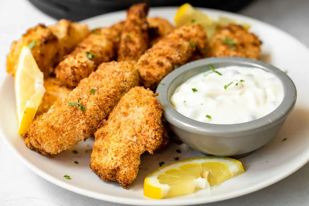 A white plate filled with golden-brown, breaded fish fillets, garnished with chopped herbs. The plate also features lemon wedges and a small bowl of creamy white dipping sauce. The background is blurred, keeping focus on the delicious meal.