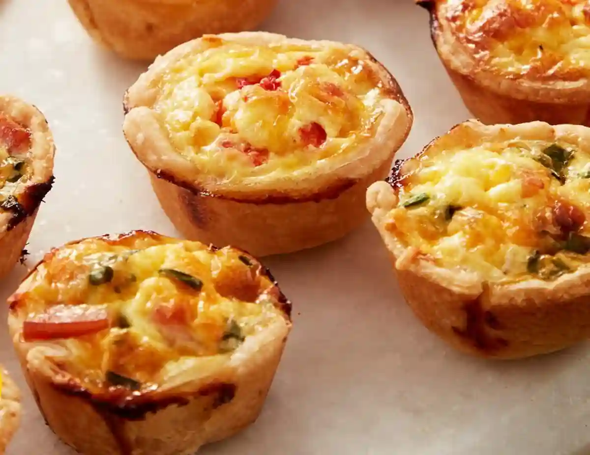 A close-up image of several mini quiches on a white surface. Each quiche has a golden-brown crust and is filled with a mixture of eggs, cheese, and diced vegetables like tomatoes and green peppers. The quiches appear freshly baked and appetizing.