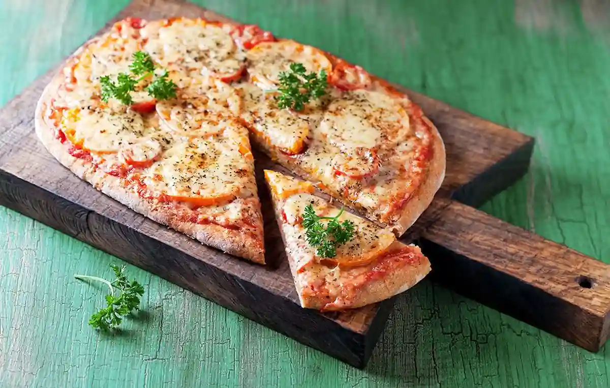 A delicious pizza on a wooden serving board. The pizza is topped with melted cheese, tomato slices, herbs, and garnished with sprigs of fresh parsley. One slice is slightly pulled out, ready to be served. The background is a rustic green wooden surface.