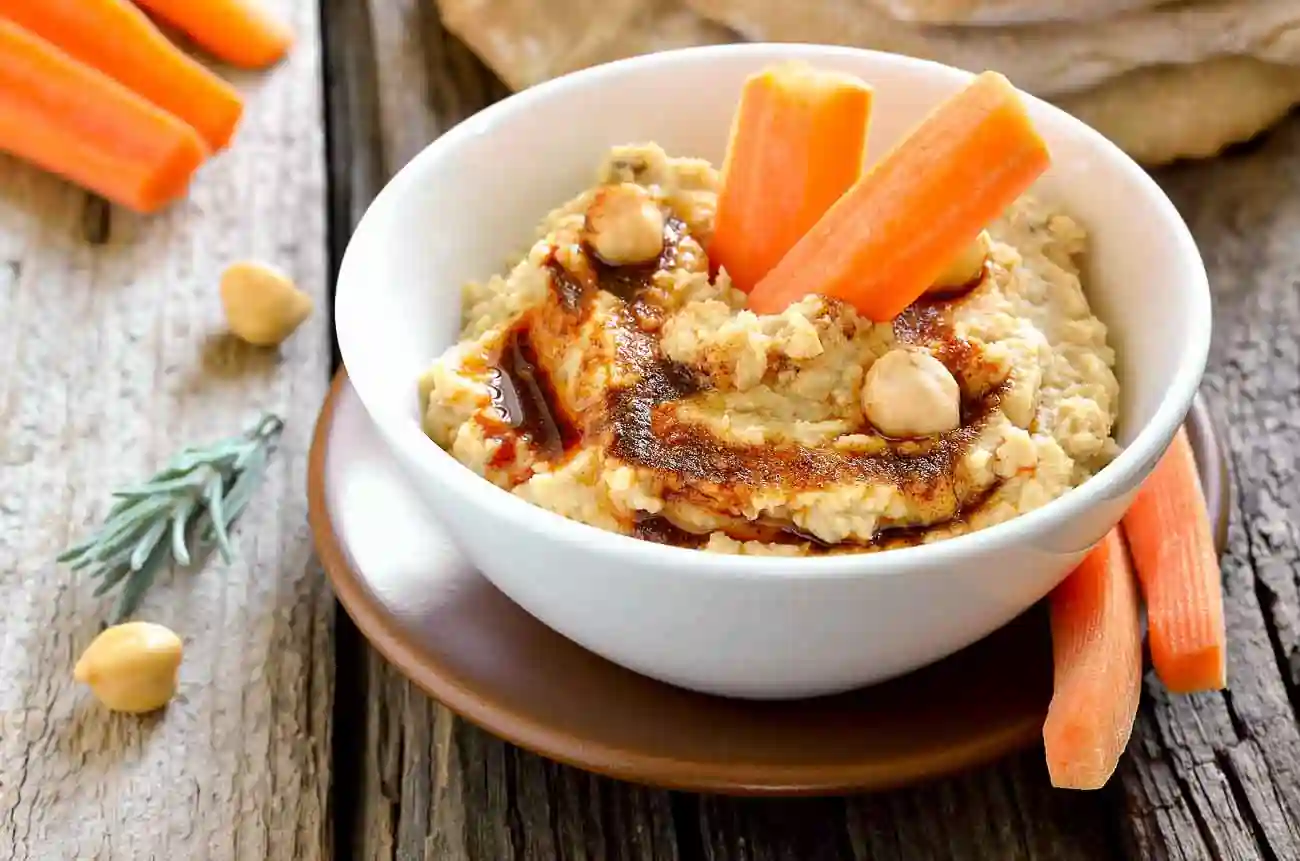 A white bowl filled with hummus, garnished with whole chickpeas, a drizzle of olive oil, and paprika. Fresh carrot sticks are placed standing in the hummus. The bowl is on a wooden table with additional carrot sticks and chickpeas scattered around.