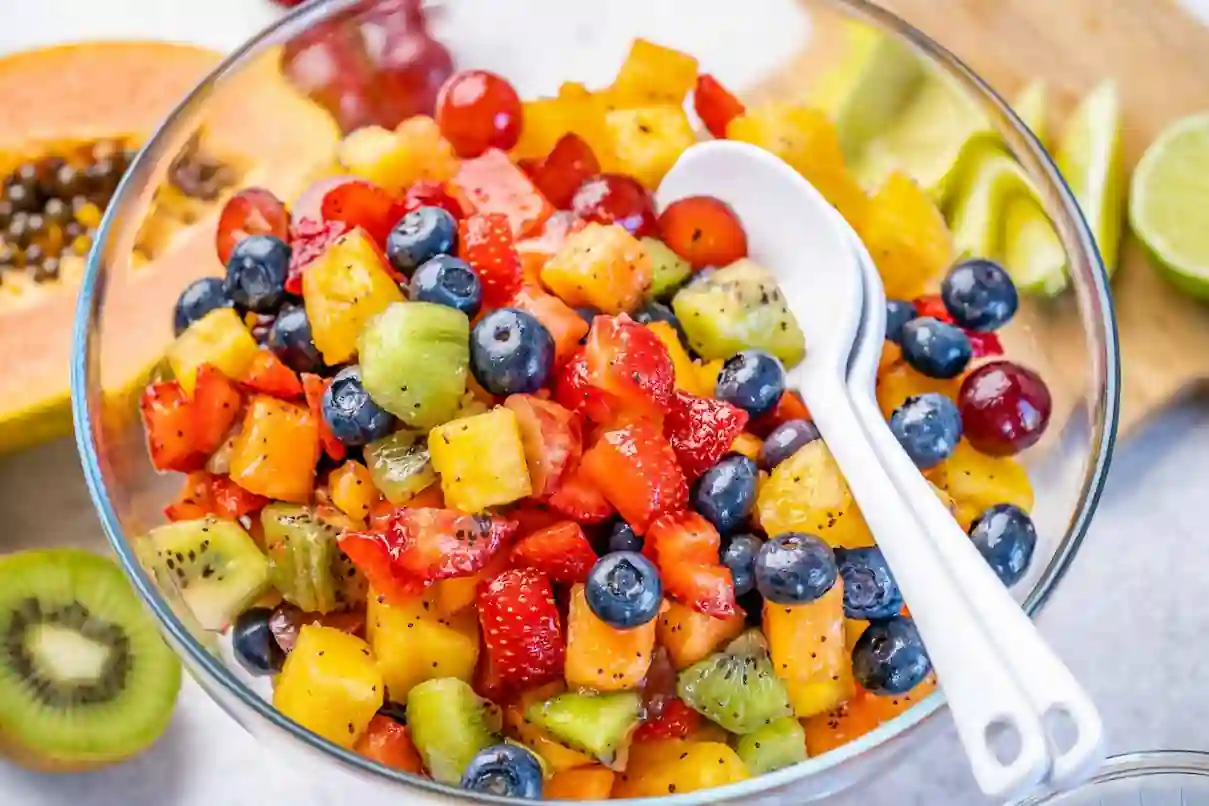 A vibrant fruit salad in a glass bowl containing a mix of diced kiwi, strawberries, blueberries, pineapple, and papaya. A white serving spoon is placed in the bowl. Sliced fruit and lime pieces are visible in the background.