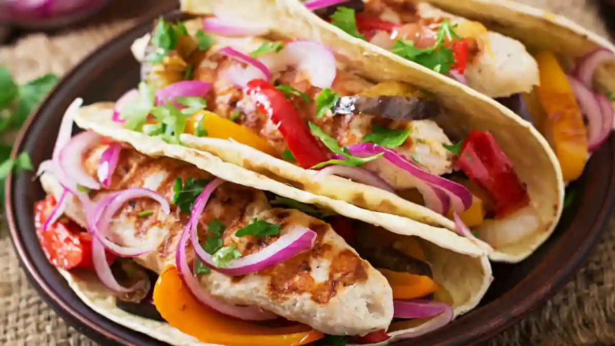 Three tacos filled with grilled chicken, sliced red onions, red and yellow bell peppers, and garnished with chopped cilantro, displayed on a dark plate on a rustic fabric background.