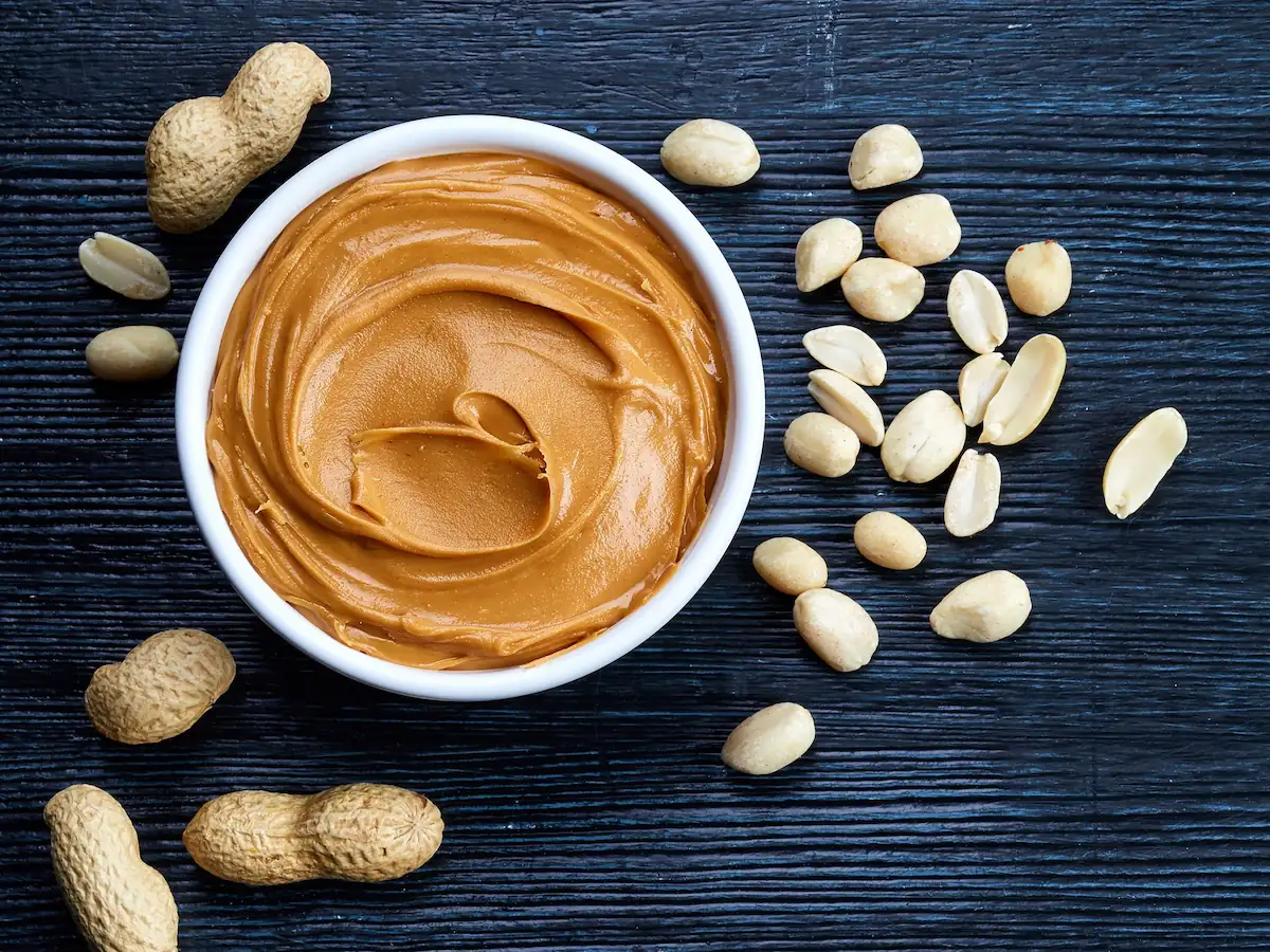 A white bowl filled with creamy peanut butter is placed on a dark wooden surface. Shelled and unshelled peanuts are scattered around the bowl. The dark background highlights the contrasting light color of the peanut butter and nuts.