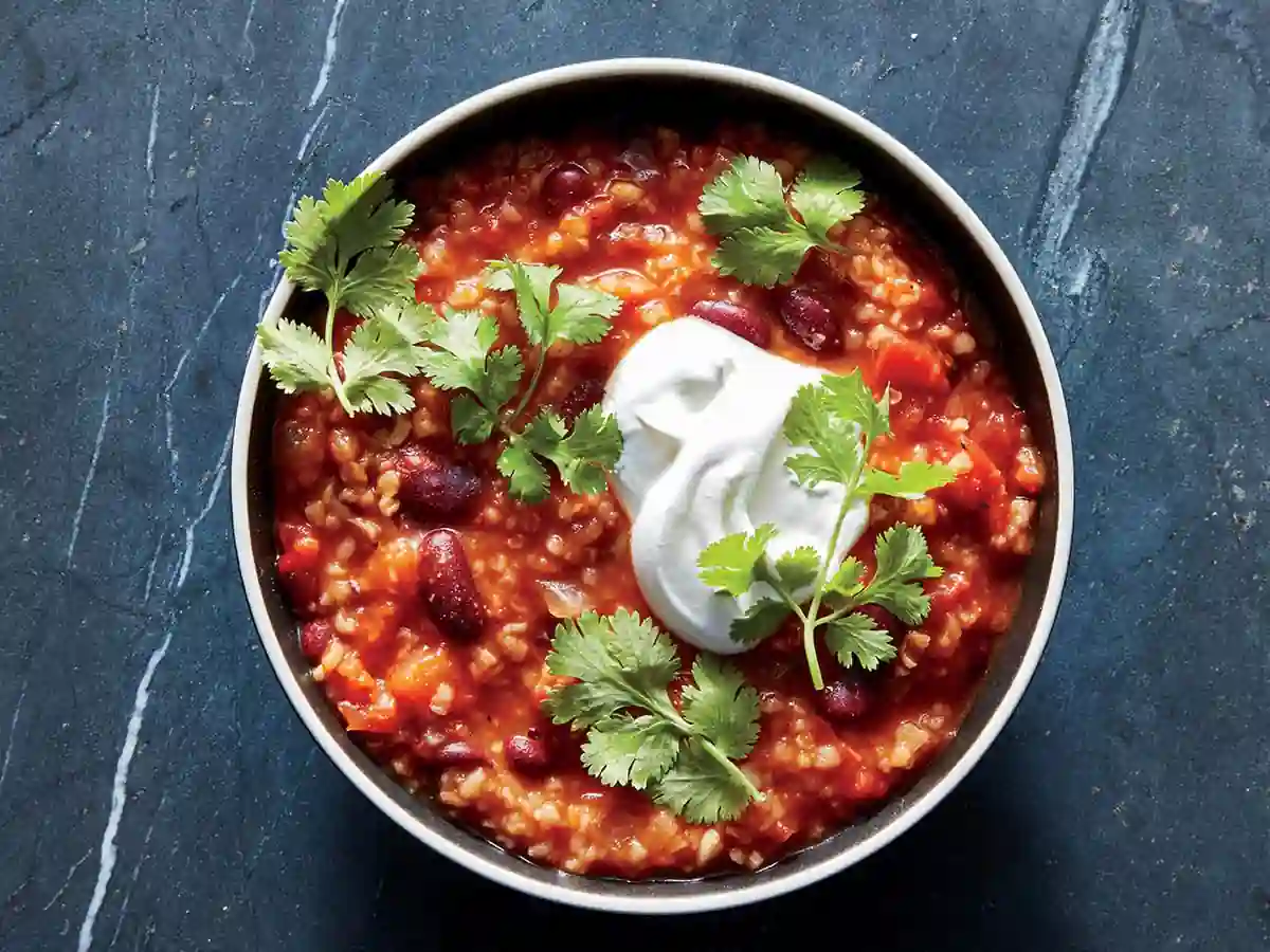 A bowl of rich, tomato-based chili topped with a dollop of sour cream and garnished with fresh cilantro leaves. The chili contains visible beans and is served on a dark stone surface.