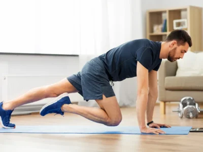 A person dressed in a navy blue shirt and shorts performs a mountain climber exercise on a blue yoga mat in a living room. Nearby, there are a couch, bookshelf, and a pair of dumbbells. Natural light enters through a window on the left.