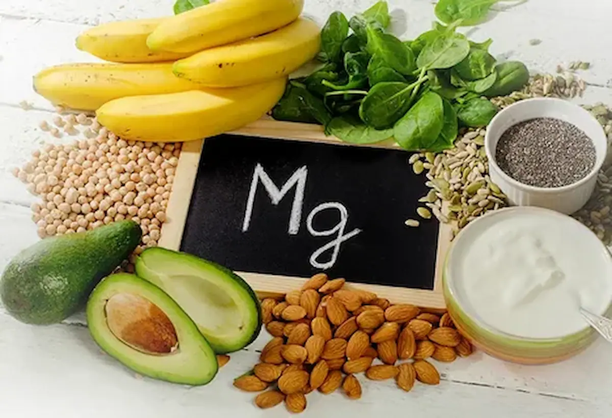 A variety of magnesium-rich foods surround a small chalkboard with "Mg" written on it. Foods include bananas, spinach, chickpeas, avocado, almonds, a bowl of yogurt, chia seeds, and sunflower seeds.