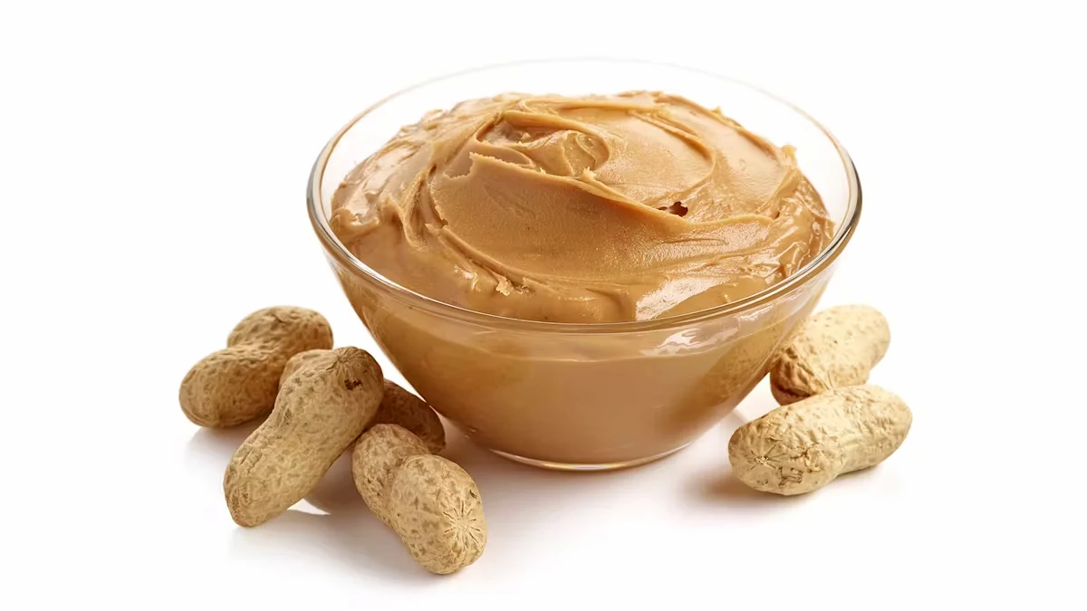 A glass bowl filled with creamy peanut butter sits beside several unshelled peanuts on a white background. The peanut butter has a smooth texture, and the peanuts are arranged around the bowl.