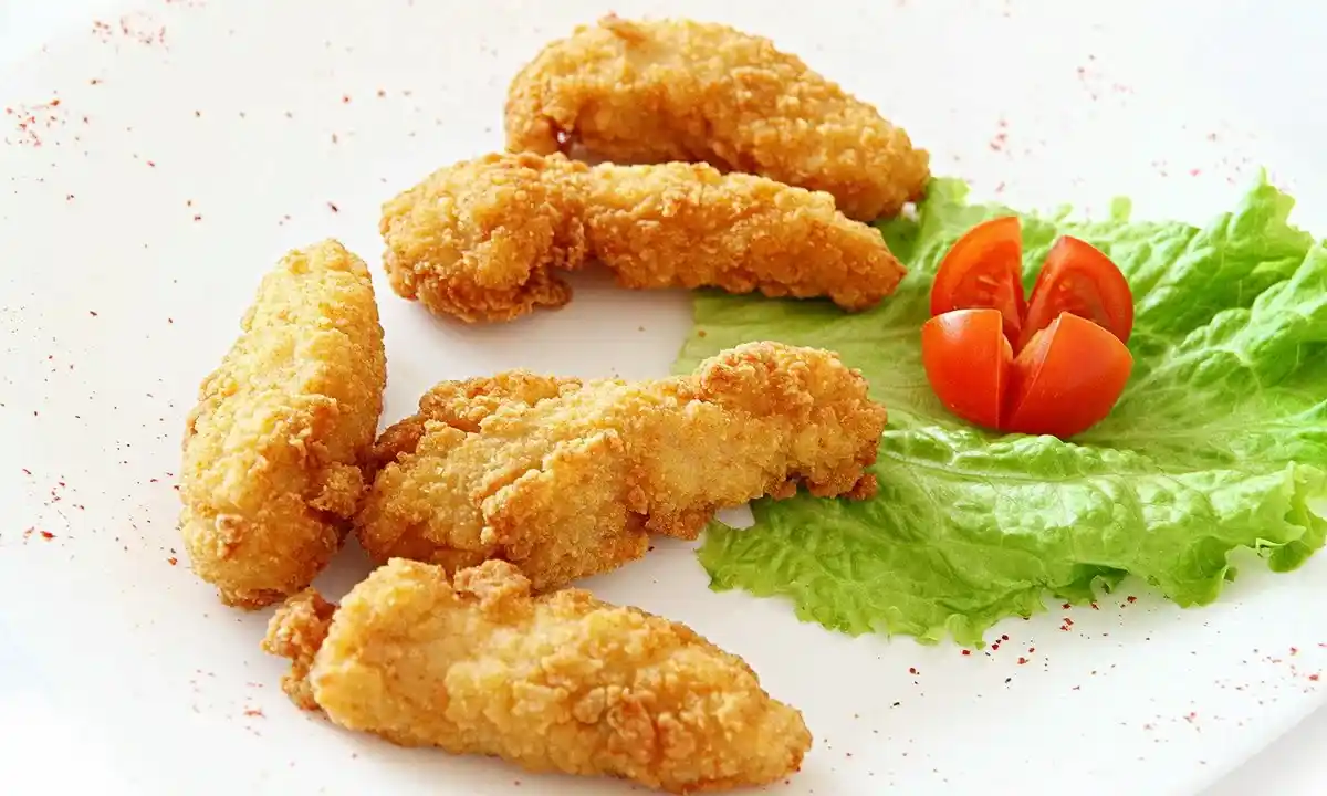 Golden brown fried chicken tenders arranged on a white plate, accompanied by a green lettuce leaf and garnished with a halved cherry tomato. A sprinkle of red seasoning is visible on the plate.