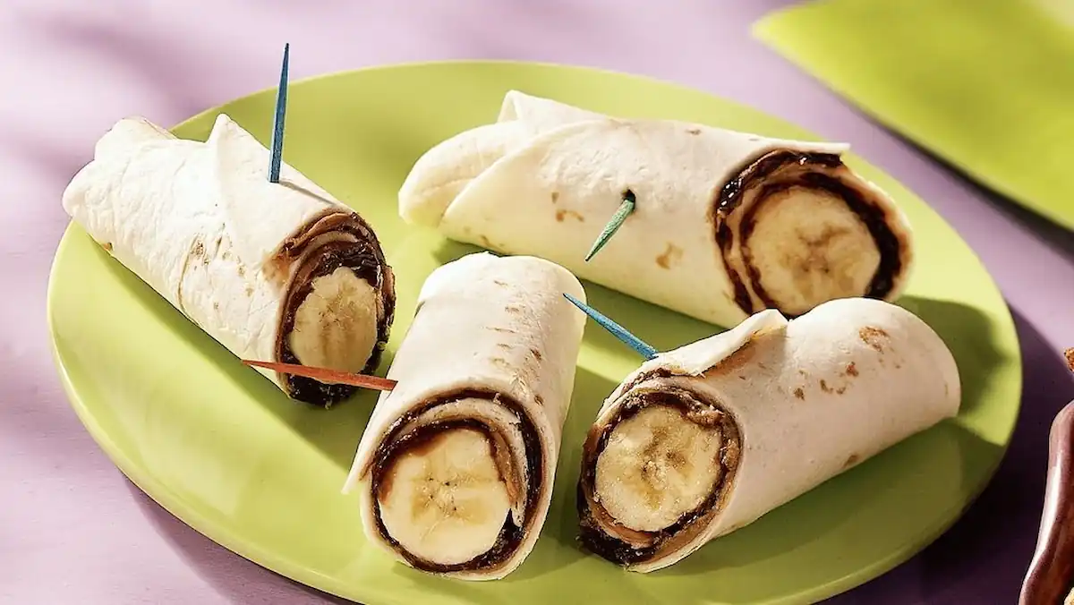 A green plate holds four banana and chocolate wraps, each secured with a colorful toothpick. The wraps are made with tortillas, filled with bananas and chocolate spread, and cut into halves. The background features a purple surface and an out-of-focus green napkin.