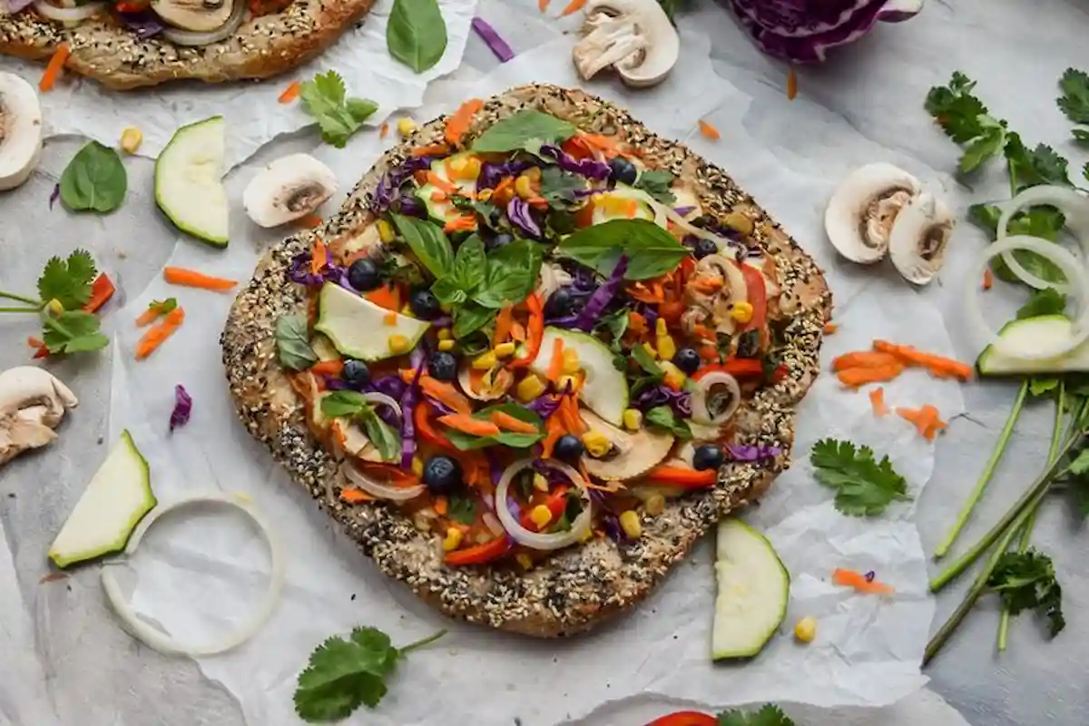 A colorful vegetable flatbread topped with slices of zucchini, onions, mushrooms, shredded carrots, purple cabbage, corn, herbs, and other vegetables. It is placed on a piece of parchment paper with additional vegetable slices and herbs scattered around.