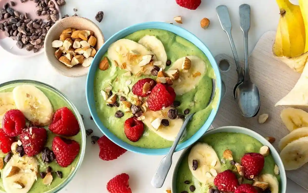 A vibrant spread featuring bowls of green smoothie topped with banana slices, raspberries, chopped nuts, and chocolate chips. Spoons are placed nearby, with additional toppings scattered around, including bananas, nuts, and chocolate chips on a light surface.