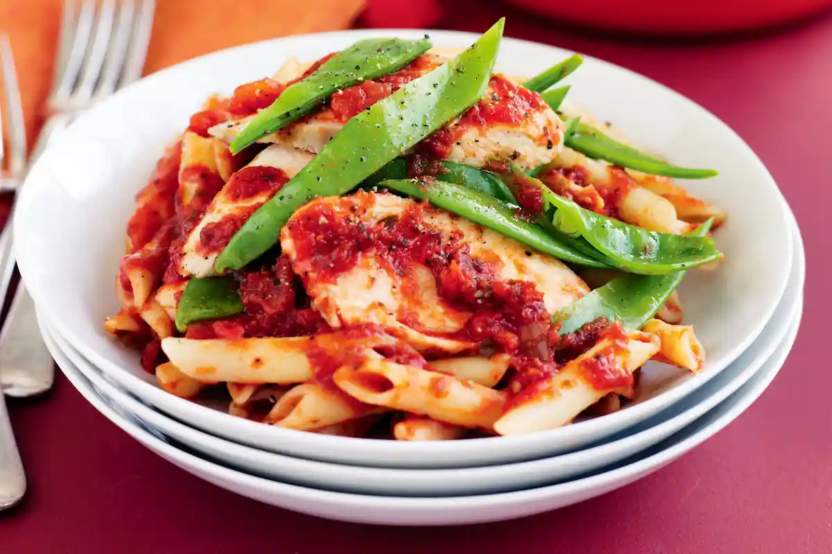 A bowl of penne pasta topped with tomato sauce, grilled chicken strips, and snow peas. The dish is garnished with black pepper. The bowl is placed on a stack of other white bowls, and there are silverware pieces on the left side.