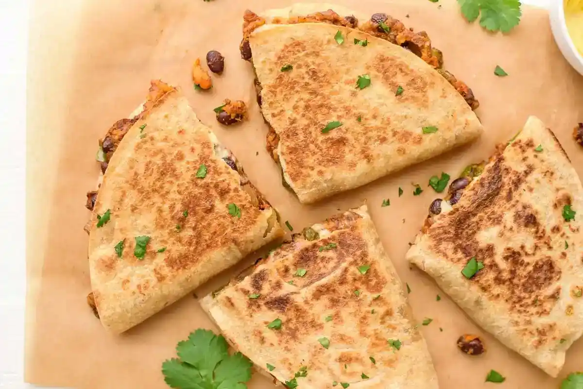 A top view of four golden-brown quesadilla wedges on a parchment paper, garnished with chopped cilantro. The filling appears to contain black beans, melted cheese, and possibly vegetables. Fresh cilantro leaves are scattered around on the parchment.