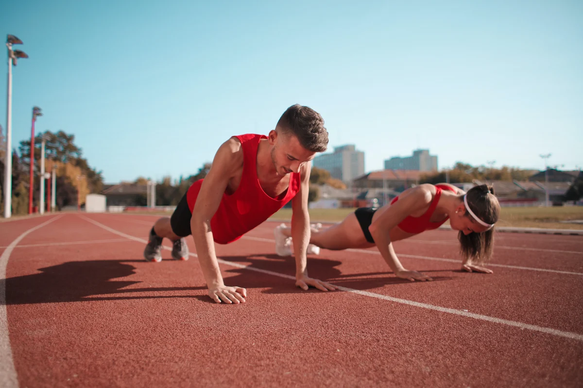 Two athletes in red workout attire performing push-ups on a running track. Both are focused, with the man on the left and the woman on the right. The background shows a clear sky, trees, and distant buildings indicating an outdoor setting.