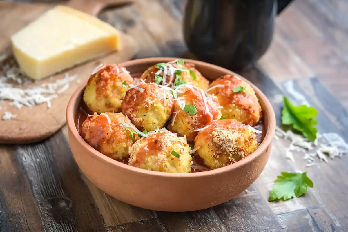 A wooden bowl filled with cheesy tomato-topped arancini sits on a rustic wooden table. Freshly grated parmesan cheese and parsley are sprinkled on top, with additional cheese and parsley scattered on the table. A piece of parmesan cheese is on a cutting board nearby.