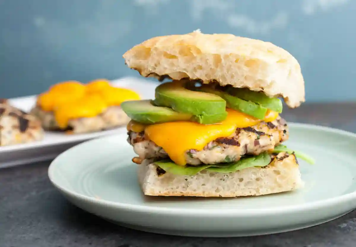 A chicken burger topped with melted cheddar cheese, fresh avocado slices, and spinach, served on a ciabatta bun with a second burger visible in the background, all on a light green plate against a blue background.