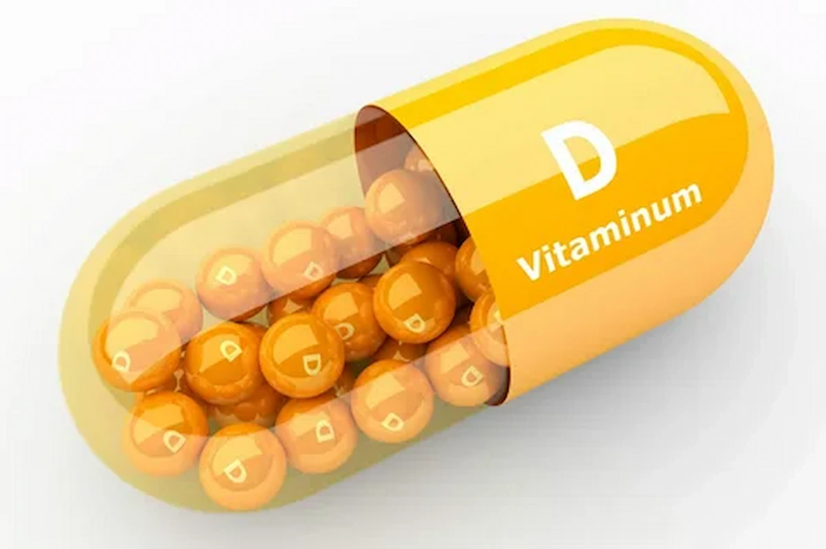 A large vitamin D capsule half-filled with smaller, round orange pellets, each marked with a "D." The capsule is partially transparent, with "Vitamin D" written on the yellow opaque part.