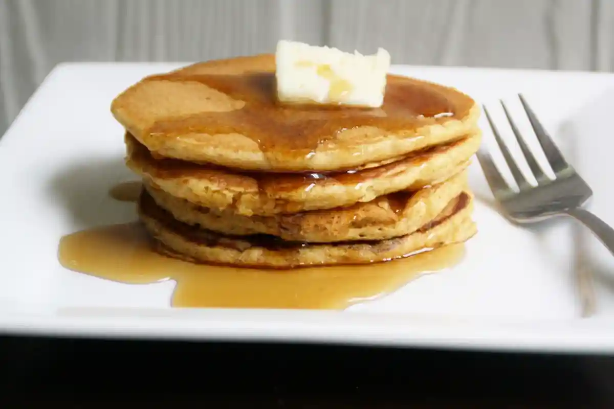 A stack of three syrup-covered pancakes topped with a pat of butter sits on a white square plate. A fork is placed on the right side of the plate. The background is out of focus.