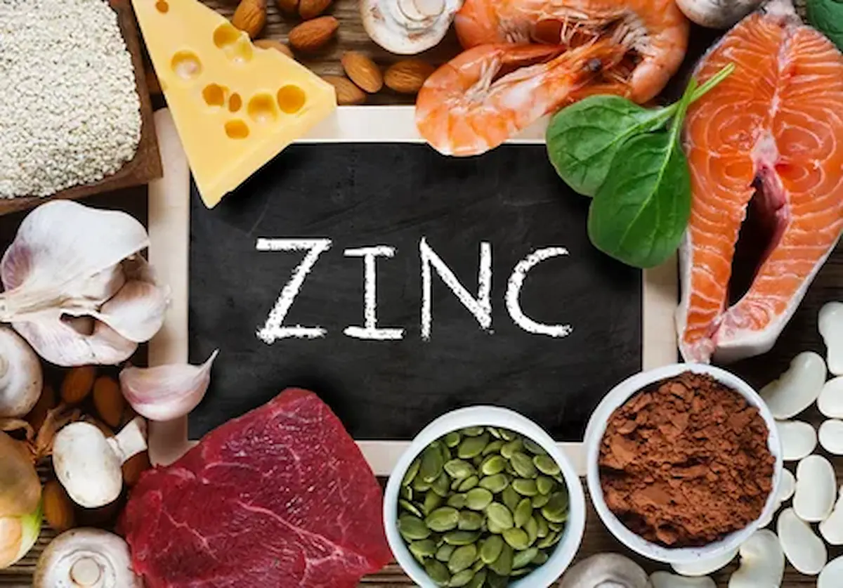 A variety of zinc-rich foods arranged around a small chalkboard with the word "ZINC" written on it. Foods include cheese, shrimp, spinach, salmon, garlic, beef, pumpkin seeds, almonds, mushrooms, sesame seeds, and cocoa powder.