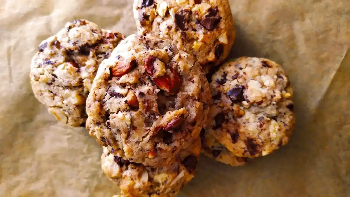 Close-up of five chunky cookies with visible chocolate chunks and nuts, placed on a sheet of brown parchment paper. The cookies have a rustic, homemade appearance with slightly uneven shapes and textures.