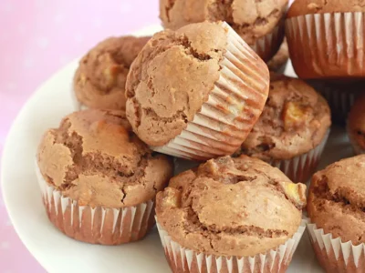 A close-up of a plate stacked with freshly baked muffins in white paper liners. The muffins are golden brown with a slightly cracked top, and they appear moist and fluffy. The background is softly blurred in pink tones.