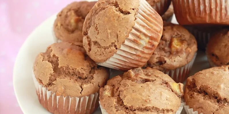 A close-up of a plate stacked with freshly baked muffins in white paper liners. The muffins are golden brown with a slightly cracked top, and they appear moist and fluffy. The background is softly blurred in pink tones.