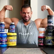 A man in a sleeveless gray "Allmax" shirt flexes his biceps while seated at a table covered with various best natural supplements for muscle growth and strength. The supplements are in containers of different sizes and colors, prominently displaying brands and labels