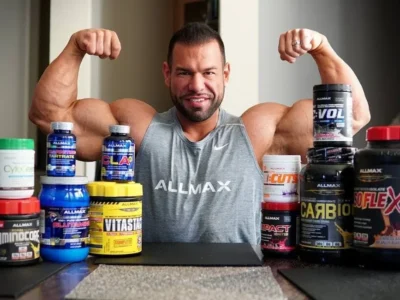 A man in a sleeveless gray "Allmax" shirt flexes his biceps while seated at a table covered with various best natural supplements for muscle growth and strength. The supplements are in containers of different sizes and colors, prominently displaying brands and labels