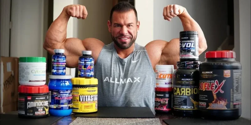 A man in a sleeveless gray "Allmax" shirt flexes his biceps while seated at a table covered with various best natural supplements for muscle growth and strength. The supplements are in containers of different sizes and colors, prominently displaying brands and labels